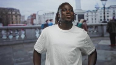 Köprü ve şehir meydanındaki anıt yanında dururken yukarı bakan ve gülümseyen bir adam rastalı beyaz t-shirt giyiyor; neşe.