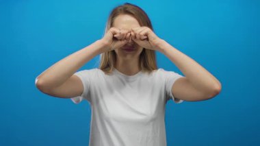 Beyaz tişörtlü beyaz bir kadın, mavi arka plan duvarına şaşkınlıkla bakıyor..