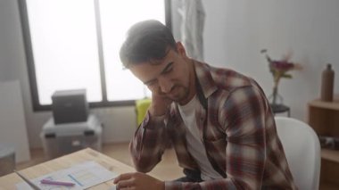 Ofisteki genç adam masadaki belgelerle dizüstü bilgisayara odaklanmış, ekose gömlek giyiyor, parlak iç mekan giyiyor, düşünceli bir ifade takınıyor, kapalı mekanda çalışıyor, modern iş ortamında çalışıyor..