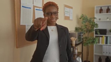 Ofisteki kamerayı işaret eden kadın, siyah ceket ve gözlük takan mantar pano çizelgeleri ve saksı bitkisiyle kamerayı işaret ediyor..