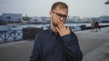 Adam, gözlük ve sakal takıp deniz kenarındaki gezinti alanında açık elle öpücük yolluyor; şakacı flört..