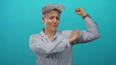 Çizgili gömlekli kadın, mavi stüdyoda pazı kasını göstermek için kolunu esnetir; kendine güvenin dayanıklılığını güçlendirir.