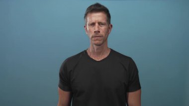 Kameraya bakan ve elini uzatan bir adam mavi arka planı ve düz siyah tişörtü olan bir stüdyoda el hareketini gösteriyor..