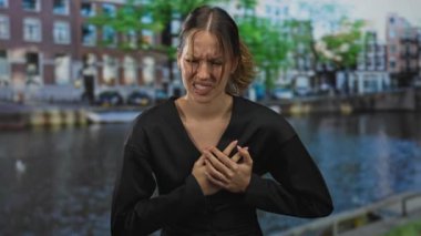 Göğüs kafesini iki eliyle tutan bir kadın, somurtkan, Amsterdam 'da kanal kenarında, tıbbi acil durum ağrısı..