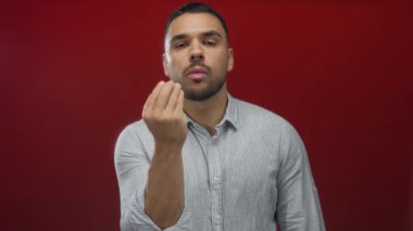 Çizgili gömlek giyen genç İspanyol adam parmak uçlarını birbirine çimdikliyor ve kırmızı stüdyo ortamında yüzün yanında ellerini kaldırıyor;.