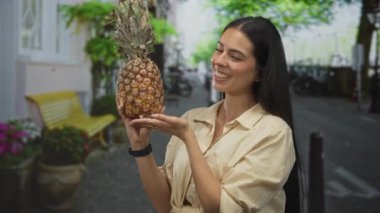 Sokakta sarı bankın ve saksı bitkilerinin yanında iki çıplak elle olgun ananas tutan kadın; taze yaz neşe atıştırmalığı.