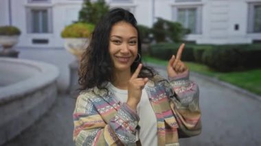 Genç İspanyol kadın açık bir yolda havuz kenarındaki çeşmeyi işaret ediyor, kameraya gülümsüyor ve göz kırpıyor; neşeli neşe.