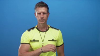 Mavi stüdyoda neon renkli sarı gömlek giyen ve boynuna ve omzuna dokunan bir iple ıslık çalan adam hakem; belirsizlik.