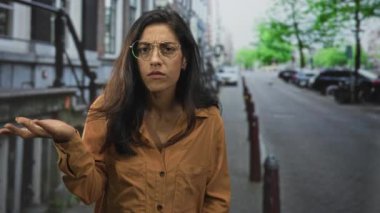 Kadın şehir caddesinde avuçlarıyla omuz silkiyor kahverengi gömlek ve gözlük takıyor, boş kaldırımda şaşkın şaşkın bakıyor; sorgulama.
