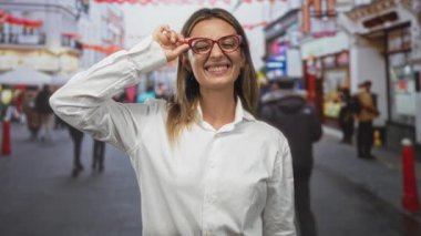 Meşgul bir şehir caddesinde kırmızı bardakları ayarlayan kadın, beyaz bir gömlek içinde gülümsüyor ve yana bakıyor; neşeli merak.