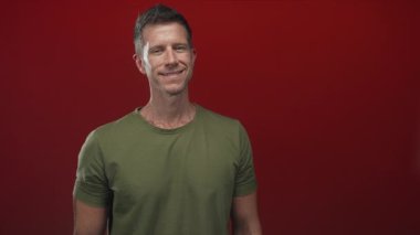 Kaya boynuzları el hareketi gösterirken gözleri kapalı gülümseyen ve stüdyoda canlı kırmızı arkaplan ile yeşil t-shirt giyen; müzik neşe dolu.