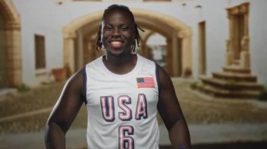 Adam işaret parmaklarını yukarı doğru işaret ediyor eski bir kasabada kaldırımlı bir sokakta, gülümsüyor ve bayrak bandı ve mavi bileklikleri olan bir USA basketbol forması giyiyor;.