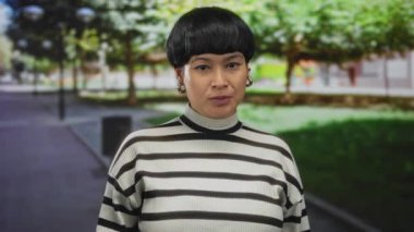 Parkta genç Çinli bir kadın. Etrafı yemyeşil, çizgili bir kazak giymiş ve yumuşak gün ışığında kendinden emin bir ifade sergiliyor..