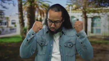 Kot ceketli ve gözlüklü bir adam gergin görünürken yeşil bir parkta palmiye ağaçları ve beyaz bir binada yumruklarını sıkıyor..