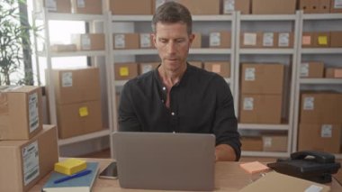 Dizüstü bilgisayarda yazan ve istiflenmiş karton kutular ve dizüstü bilgisayarlar arasında akıllı telefonu kontrol eden bir adam..