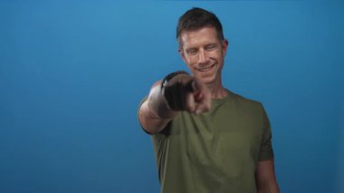 Stüdyoda elini kameraya doğrultup gülümseyen ve kalp hareketi yapan bir adam..