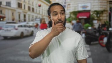 Kalabalık bir caddede, arabaların ve scooterların arasında el hareketleriyle göğsüne dokunan beyaz tişörtlü sakallı adam düşünceli..