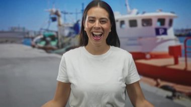 Genç esmer kadın, iskelede beyaz tişört ve ukala giyinmiş bir balıkçı teknesinin yanında el çırpıyor ve gülümsüyor..