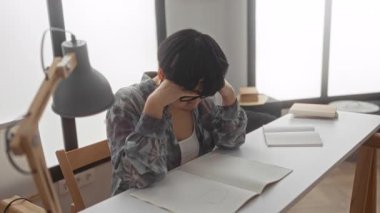 Bir dairede oturan genç bir kadın elinde lamba ve notlarla bir masada ders çalışıyor. Gözlüklerini düzeltirken stresli görünüyor..