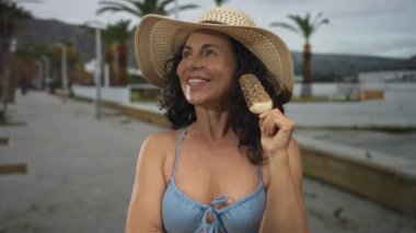 Orta yaşlı, bikinili bir kadın sokakta palmiye ağaçlarıyla dondurma yiyor, şapkası var ve güneşli bir günde neşeyle gülümsüyor..