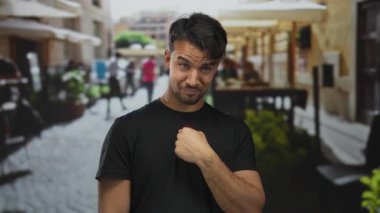 Genç ispanyol adam, parmakları dışarıda, teras kafesinin arka planında iken el kol hareketi yapıyor.