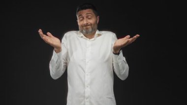 Adam açık ellerle omuz silkiyor ve siyah bir stüdyoda omuzlarını kaldırıyor, günlük beyaz gömlek ve budanmış sakal, sorgulama ifadesi; belirsizlik.