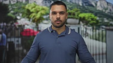 Mavi polo giyen adam demir parmaklıkların yanında göğsünü işaret ediyor. Açık hava sokak ortamında tırabzan kullanıyor. Kendine güven..