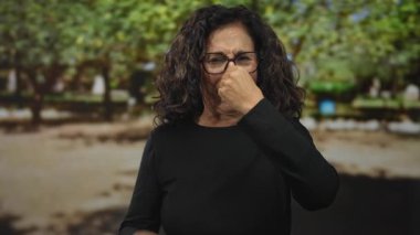 Parkta burnunu tutan bir kadın açık havada güneşli bir günde arka planda yeşil ağaçlardan tiksindiğini ifade ediyor..