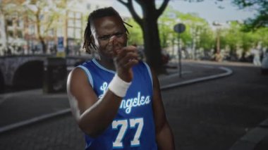 Mavi Los Angeles basketbol forması giyen bir adam, şehrin bir caddesinde, bir kanal köprüsü ve ağaçların yanında çimdikleme jestinde kameranın yanında el ele tutuşuyor..