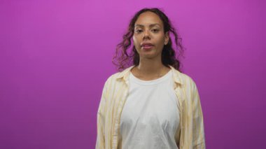 Kadın, beyaz tişört ve sarı çizgili tişört giyen, mor stüdyoda görünür parmak hareketleriyle kamerayı işaret ediyor..