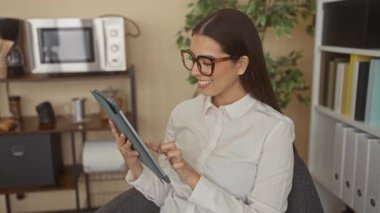 Ofis sandalyesinde parmak ile tablet ekranı tıklatan kadın, gözlük takarken gülümseyen ve raflar ve bitki yakınında beyaz bir bluz giyen; üretkenliğe odaklan.