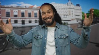 Sakallı, gülümseyen ve elleri açık bir adam kot ceket ve beyaz tişört giyen güneşli bir sokakta park edilmiş bisiklet ile; neşeli bir karşılama.