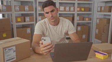 Akıllı telefon tutan ve dizüstü bilgisayar ekranını kontrol eden bir adam, bir teslimat bürosu binasında raflarda istiflenmiş kutular arasında; konsantrasyon.