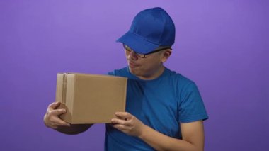 Mor bir stüdyoda mavi başlık, mavi gömlek ve gözlük takarken göğsünde mühürlü bir karton koli tutan genç kurye..