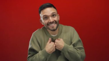 Genç adam enerjik bir şekilde kırmızı bir zemine karşı gülümsüyor sıcak bir kazak giyiyor ve mutluluk ve heyecan ifade ediyor..