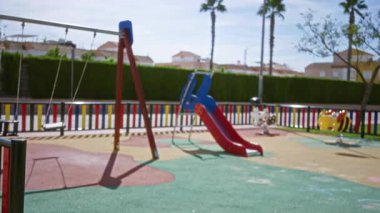 Oyun parkı odaklı olmayan kaydırak çitleri ve renkli kauçuk yüzey dışarıda; arka plan telif alanı sakin.