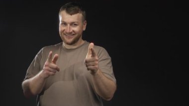 Siyah stüdyoda gülümseyen ve parmaklarını kameraya gösteren adam, çıplak eller görünür ve sıradan tişörtler; oyuncu tavırlar..