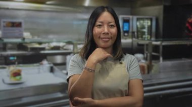 Önlüklü, eli çenesinde ve ön kolu görünen bir kadın. Mutfak binasında duruyor. Düşünceli bir kararlılık..