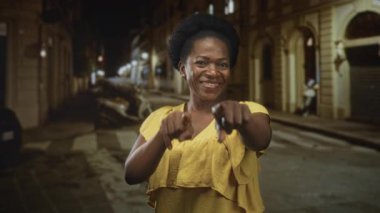 Sarı elbiseli kadın gece loş bir şehir caddesinde el kol hareketi yaparak kameraya doğru gülümsüyor, neşeli neşe saçıyor..