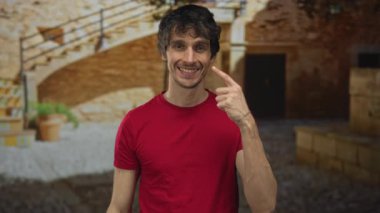 Kırmızı gömlekli adam tuğla merdivenlerin yanındaki tarihi şehir sokağında parmak yanağa parmakla işaret ediyor..