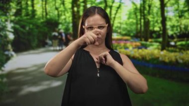 Kadın kamerayı işaret eder ve orman yolunda gözlük ve siyah kolsuz bluz giyerek önünde yumruğunu bastırır; kararlılık.