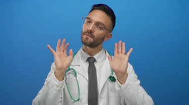 İspanyol erkek doktor gözlüklü ve steteskoplu beyaz önlüklü. Mavi arka planda sükunet için el kol hareketi yapıyor..