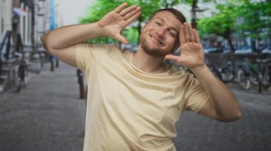 Bej t-shirt ile kaldırımlı sokakta yüzü çerçeveli adam görünür sakallı geniş gülümseme; oyuncu güven.