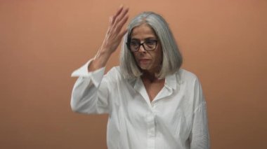 Beyaz saçlı bir kadın stüdyoda şeftali rengi arka plan ve beyaz gömlekle el ele tutuşuyor..