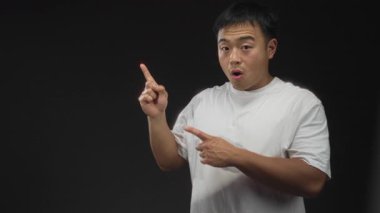 Beyaz tişörtlü genç Çinli adam her iki işaret parmağını da stüdyoda düz siyah arkaplan ile boş alana işaret ediyor.