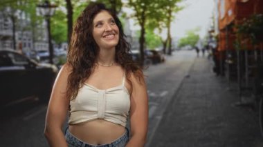 Kadın, ağaçlarla ve park halindeki arabalarla dolu bir şehir sokağına bakarken göbek atar ve gülümser; neşeli iyimserlik.