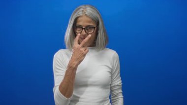 Kadın, canlı mavi bir stüdyo duvarına karşı eliyle barış işareti gösteriyor, gözlük ve beyaz bluz giyiyor, dinginlik iyimserliği bilgeliği.