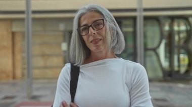 Gözlük takan gri saçlı kadın, şehir caddesinde omuz çantası askısı taşıyan orta parmağını gösteriyor..