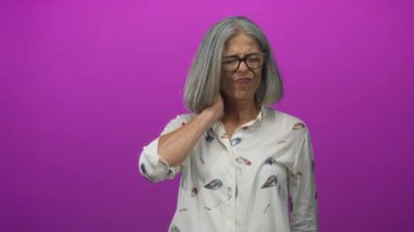 Mavi duvara karşı boynu bağlı bir kadın, gözlük ve çiçekli bluz giyiyor; rahatsız edici sertlik.