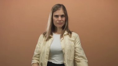 Sarışın genç kadın, turuncu stüdyoda hem beyaz atlet hem de bej gömlek giyen orta parmak hareketlerini sergiliyor..